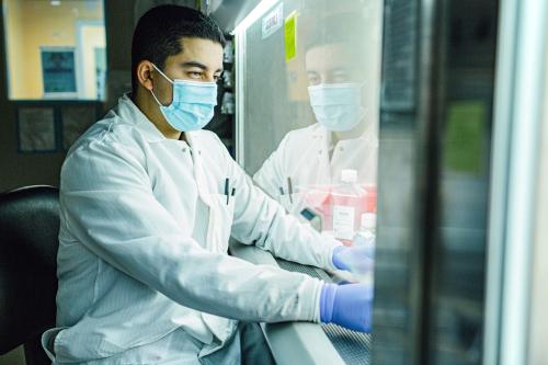 A masked UCLA researcher sitting in front of hooded lab equipment and pipetting 